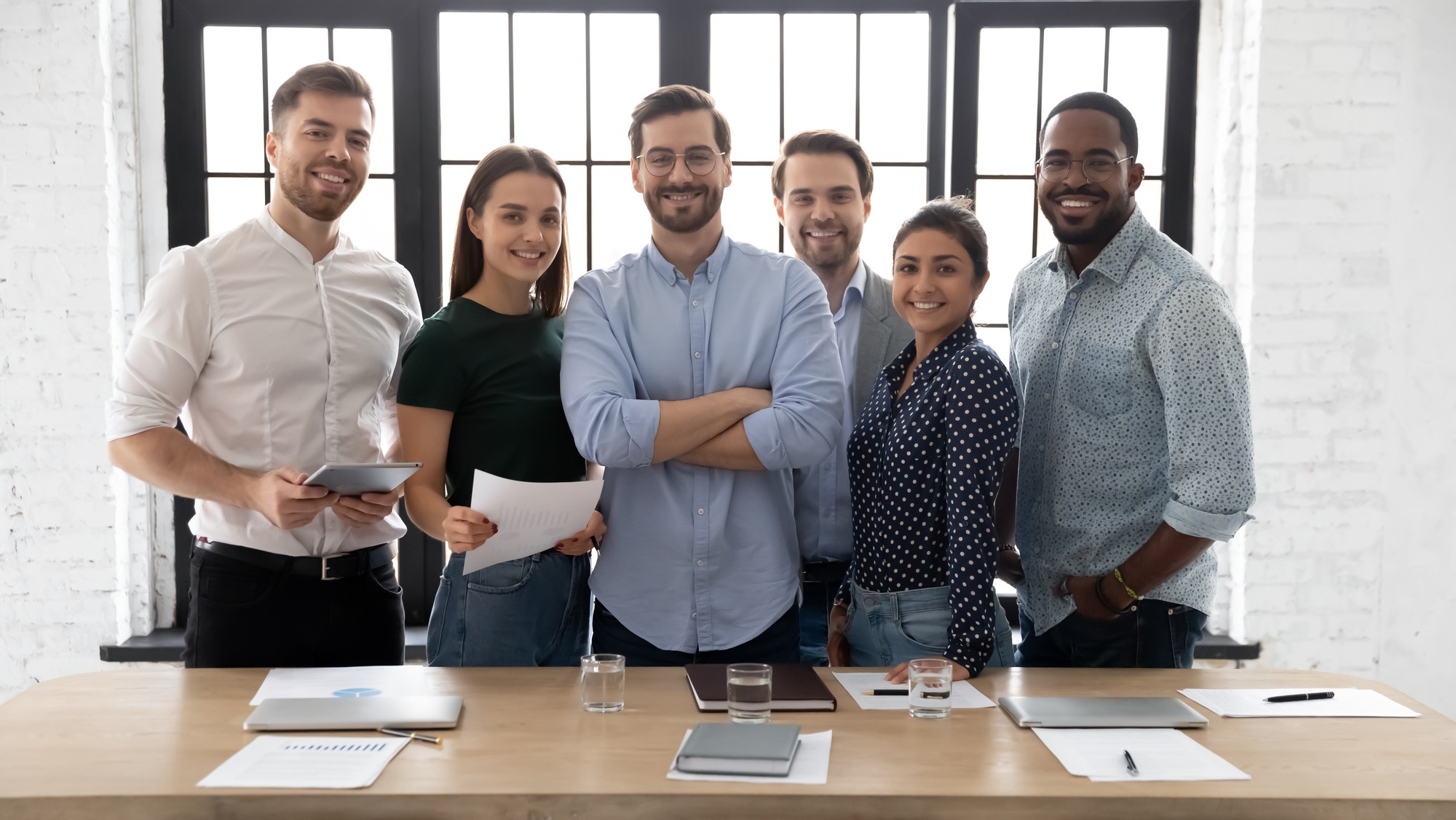 A diverse team of six professionals smiles confidently in a bright office setting, holding documents and tablets, suggesting collaboration and teamwork.